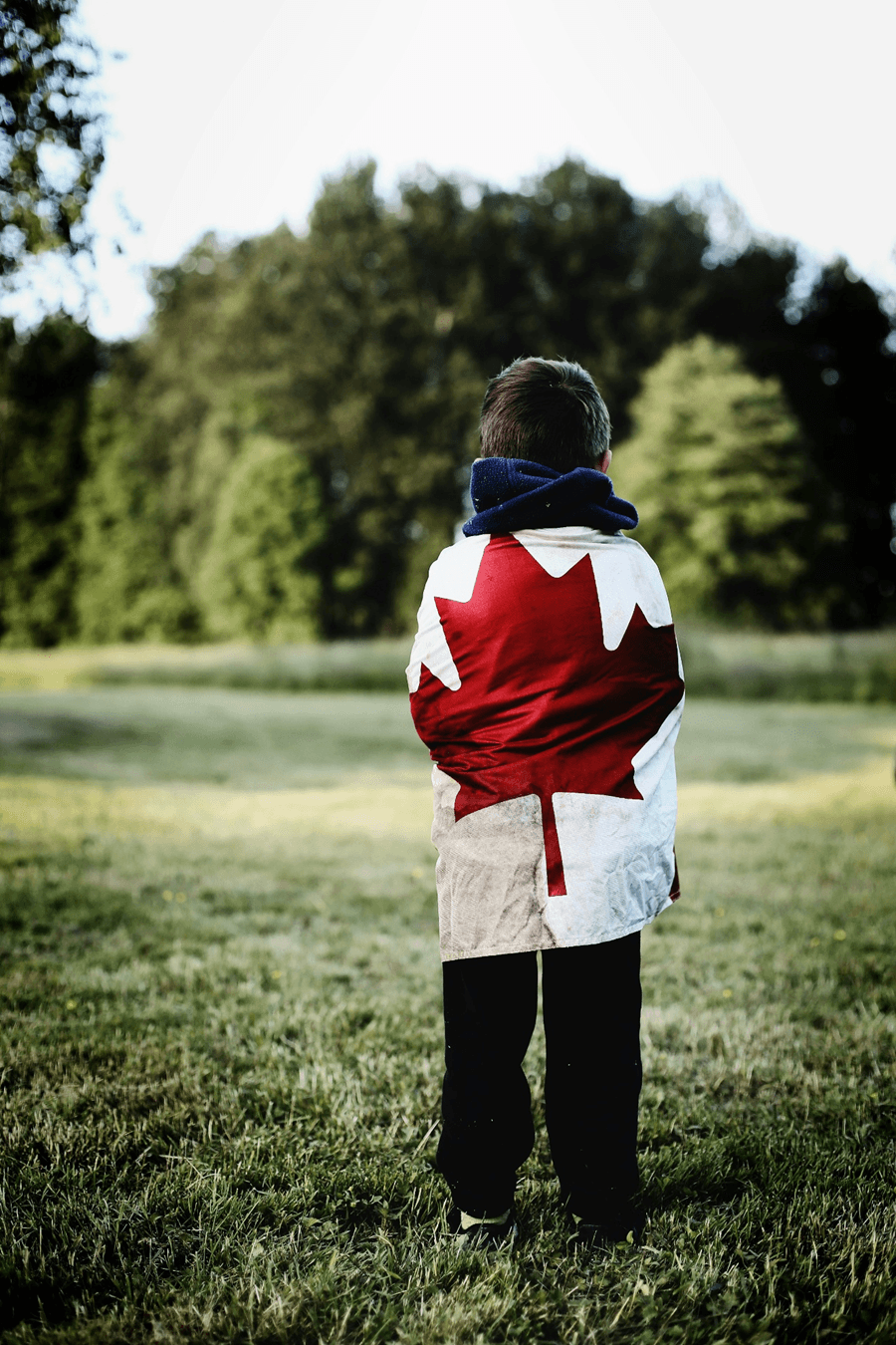 A young boy stands outdoors wrapped in a Canadian flag, suggesting Canada Day celebration in Calgary, Alberta.